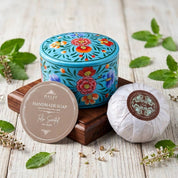 A round turquoise tin of handmade soap with colorful floral patterns sits on a wooden block beside a wrapped soap bar and a round lid label reading 'FAMILY Handmade Soap The Sweet.' Green leaves and small dried flowers are scattered on a whitewashed wood surface.