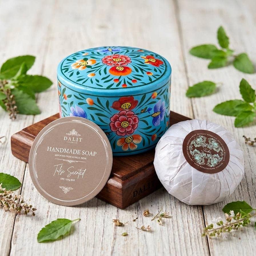 A round turquoise tin of handmade soap with colorful floral patterns sits on a wooden block beside a wrapped soap bar and a round lid label reading 'FAMILY Handmade Soap The Sweet.' Green leaves and small dried flowers are scattered on a whitewashed wood surface.