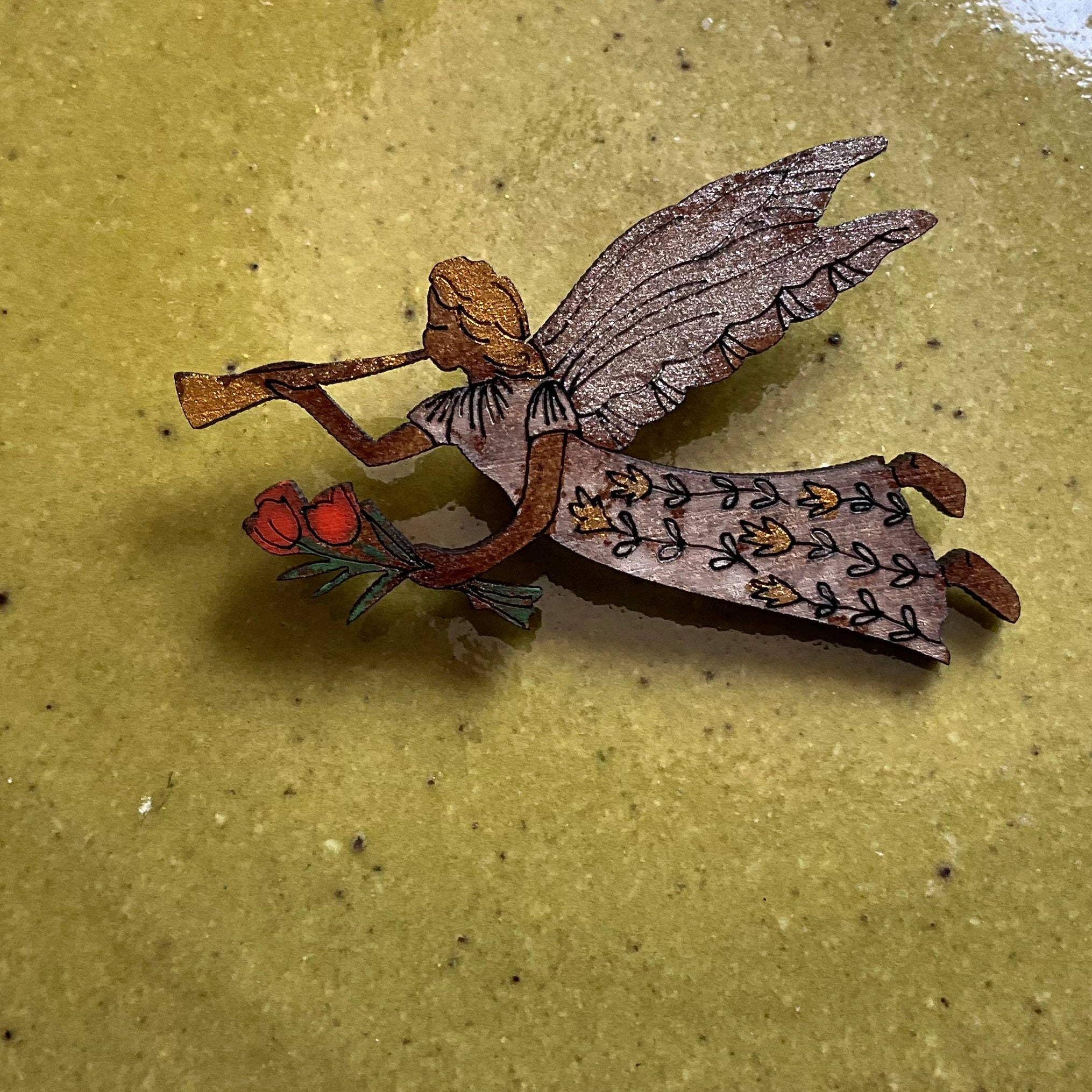 A small hand-painted wooden fairy figurine with wings and a patterned dress, holding red berries, resting on a textured olive-green surface.