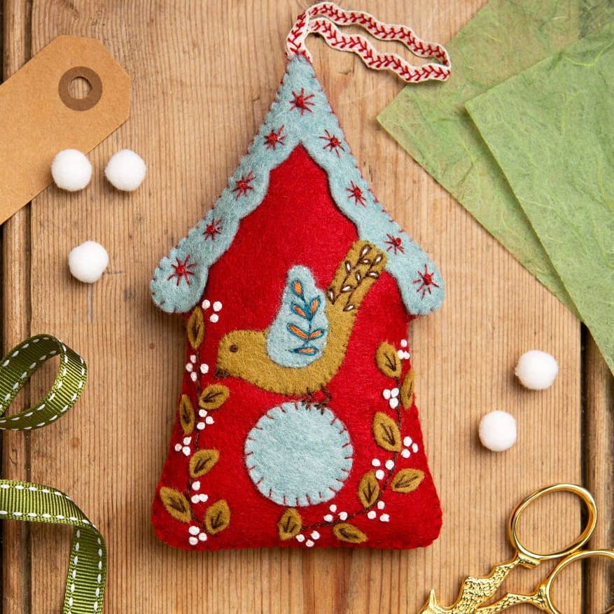 A handmade red felt ornament shaped like a house with a blue roof, embroidered bird, and white flowers, displayed on a wooden surface with ribbon, scissors, pom-poms, and green paper.