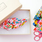 A knotted fabric bracelet with a colorful floral print in pink, red, green, and blue, presented in a light beige box with a silver-toned metal clasp. The box lid has 'Jenny Bosworth' printed on it, and sewing-themed props like scissors and fabric are visible on the right.