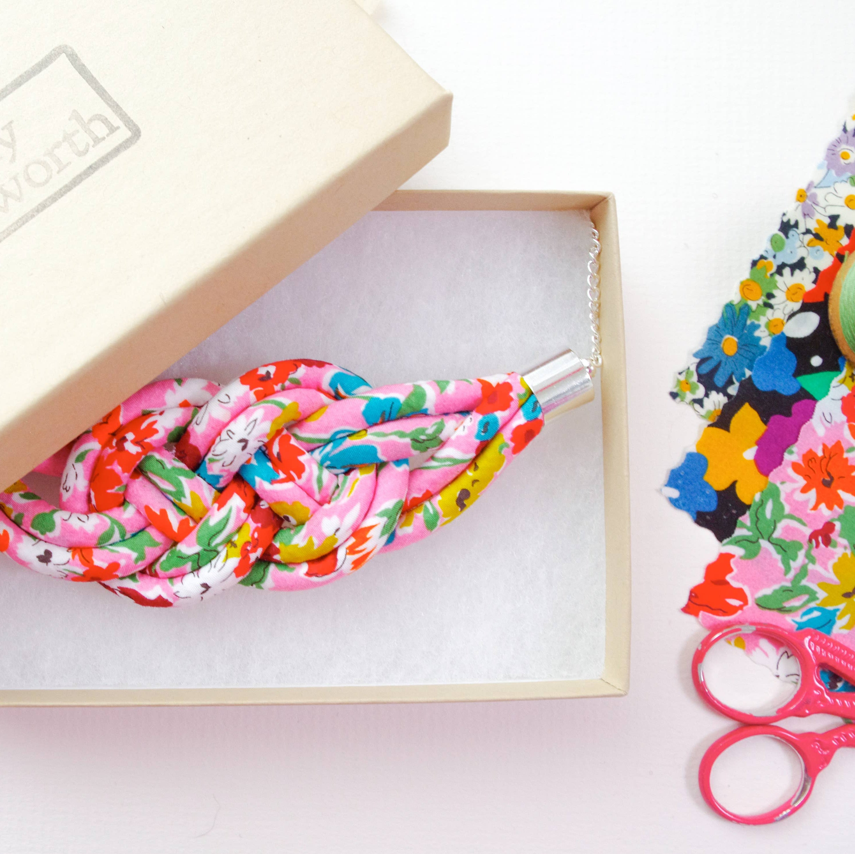 A knotted fabric bracelet with a colorful floral print in pink, red, green, and blue, presented in a light beige box with a silver-toned metal clasp. The box lid has 'Jenny Bosworth' printed on it, and sewing-themed props like scissors and fabric are visible on the right.