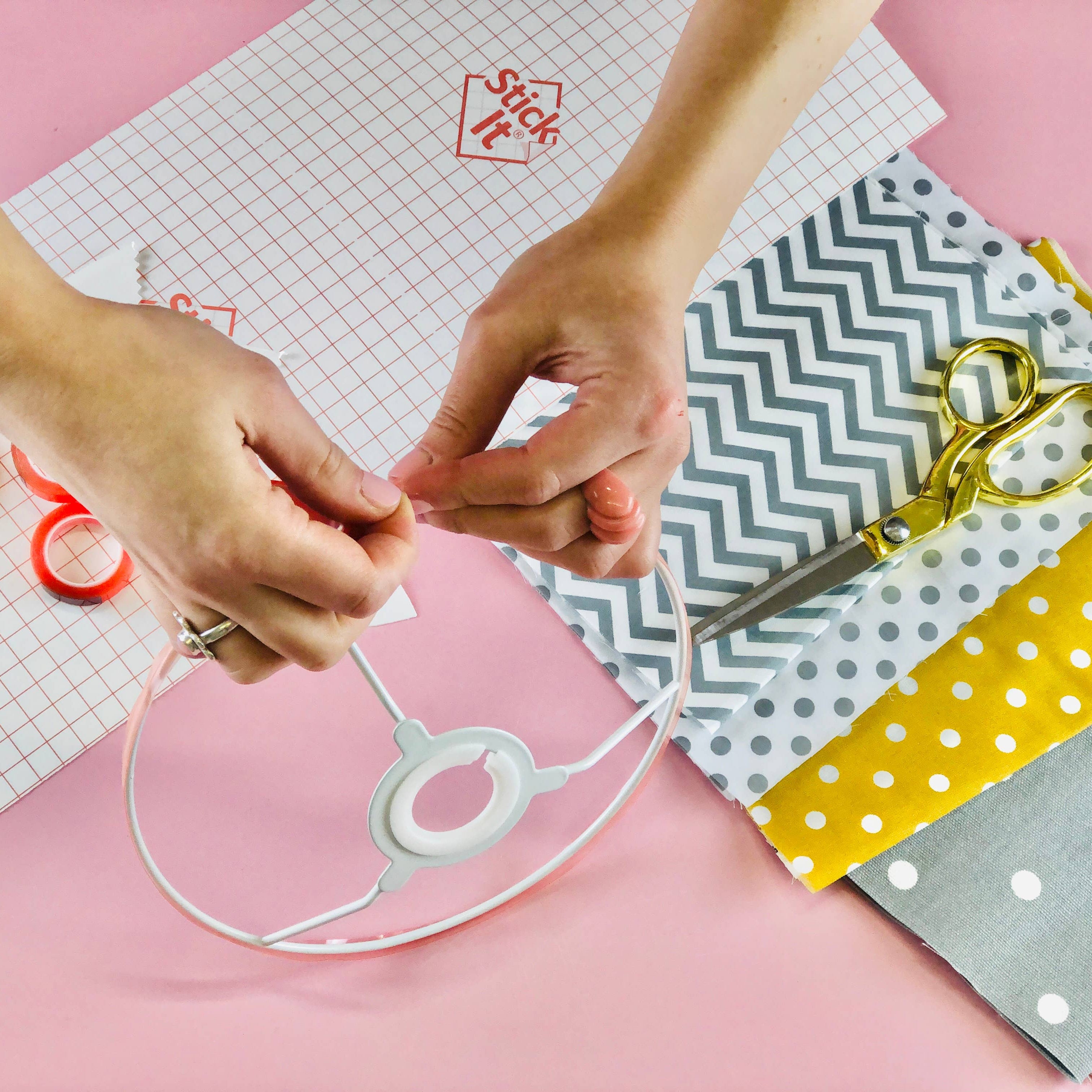 A person's hands are shown applying a clear adhesive patch to a white circular plastic ring, set against a pink background with craft supplies including patterned papers, scissors, and tape.