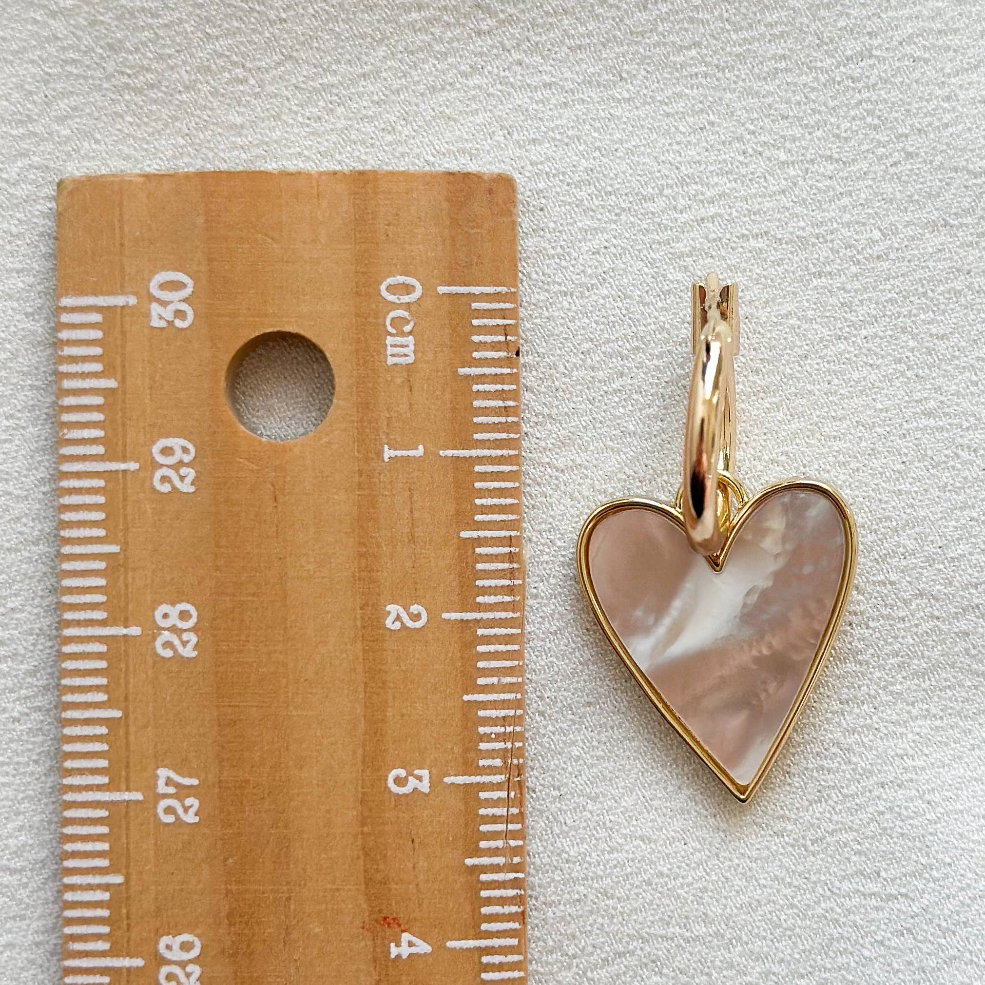 A small heart-shaped pendant with a gold-tone border and a pinkish mother-of-pearl inlay, shown next to a wooden ruler on a white textured fabric background.