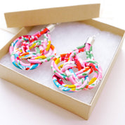 A pair of colorful woven fabric earrings displayed inside an open kraft gift box with white foam insert on a white background.