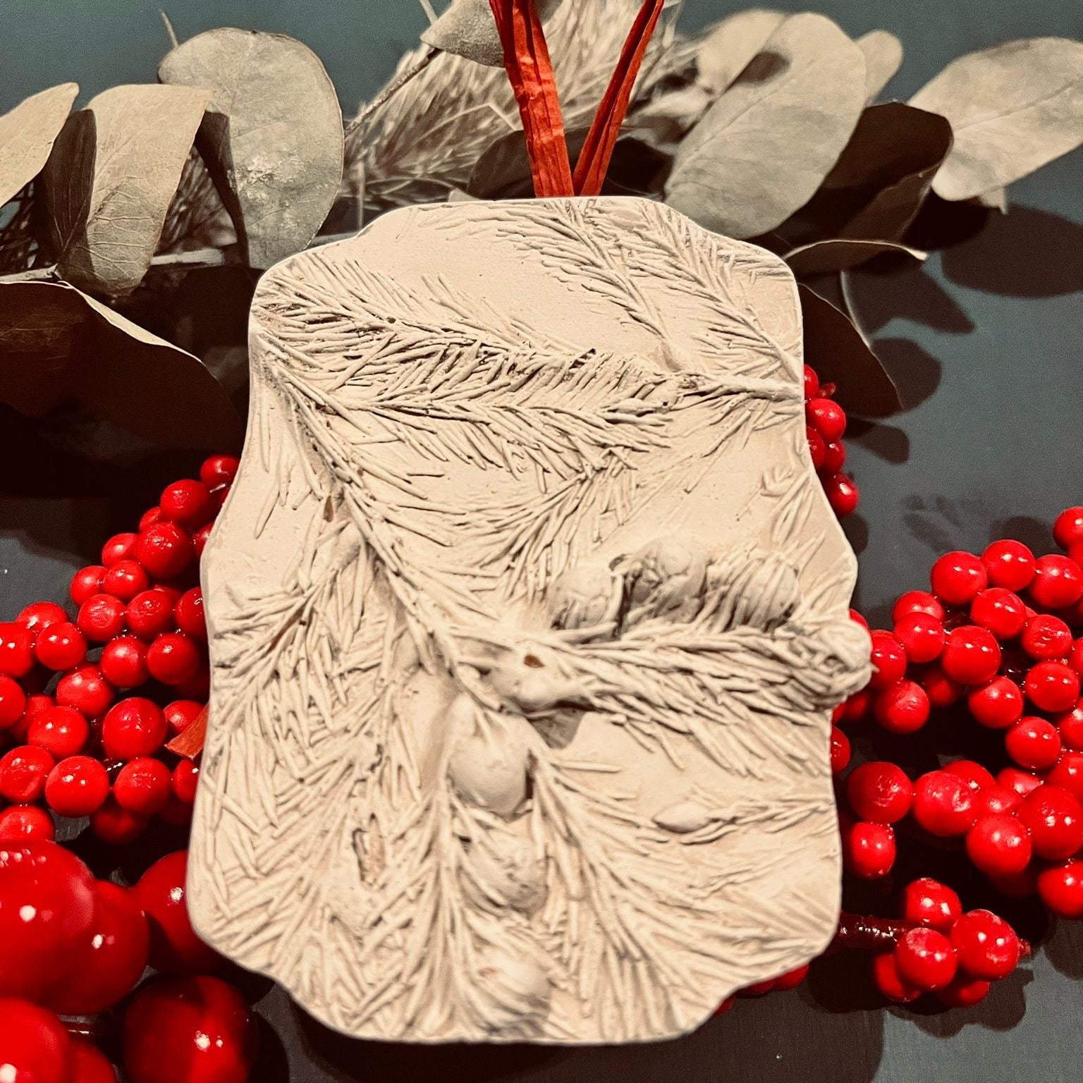 Textured beige object with red ribbon on a dark surface with red berries and gray leaves.