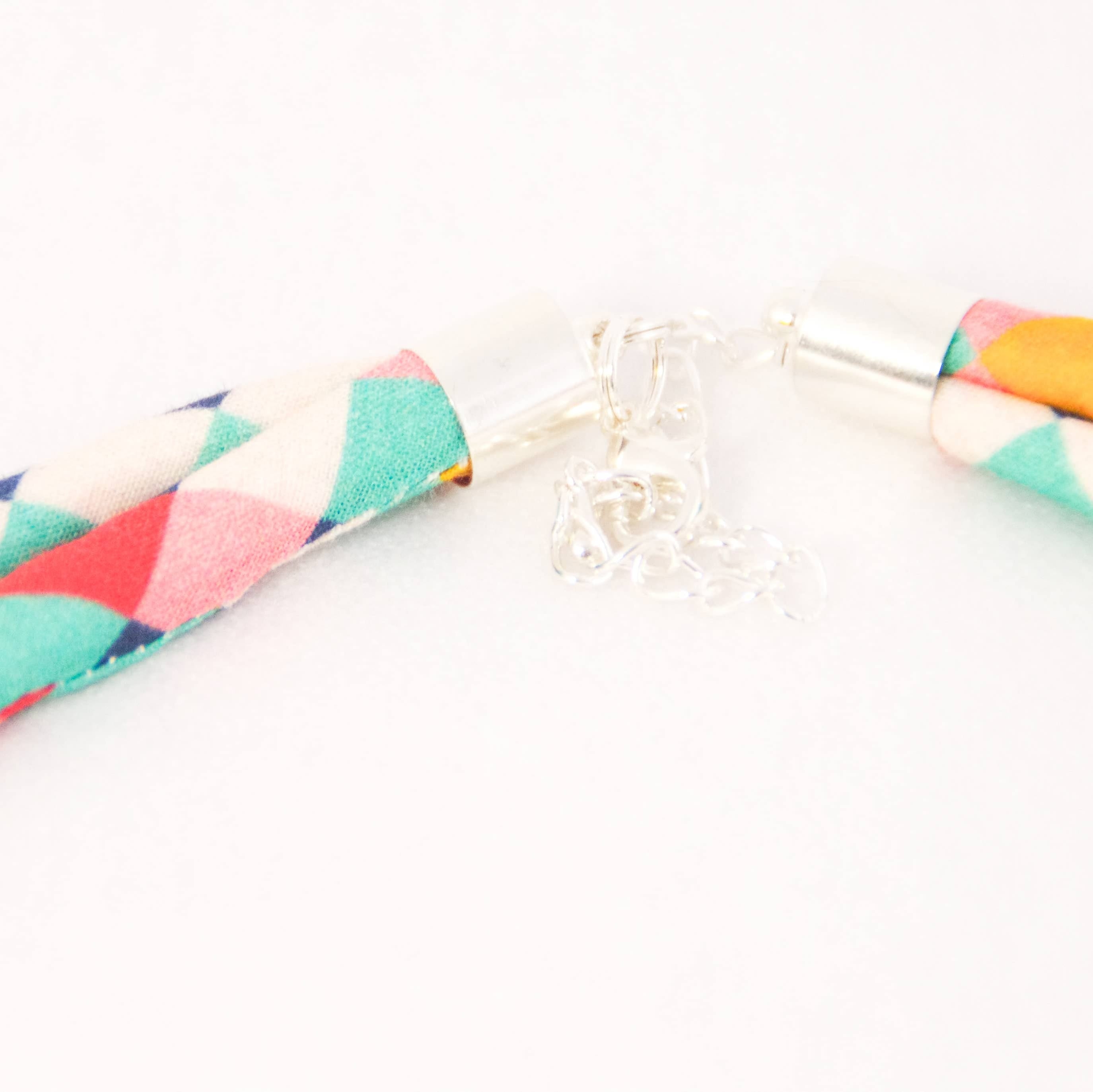 A close-up of a braided fabric necklace with a colorful geometric pattern of pink, teal, orange, and white triangles, finished with silver metal caps and a small silver charm, on a white background.