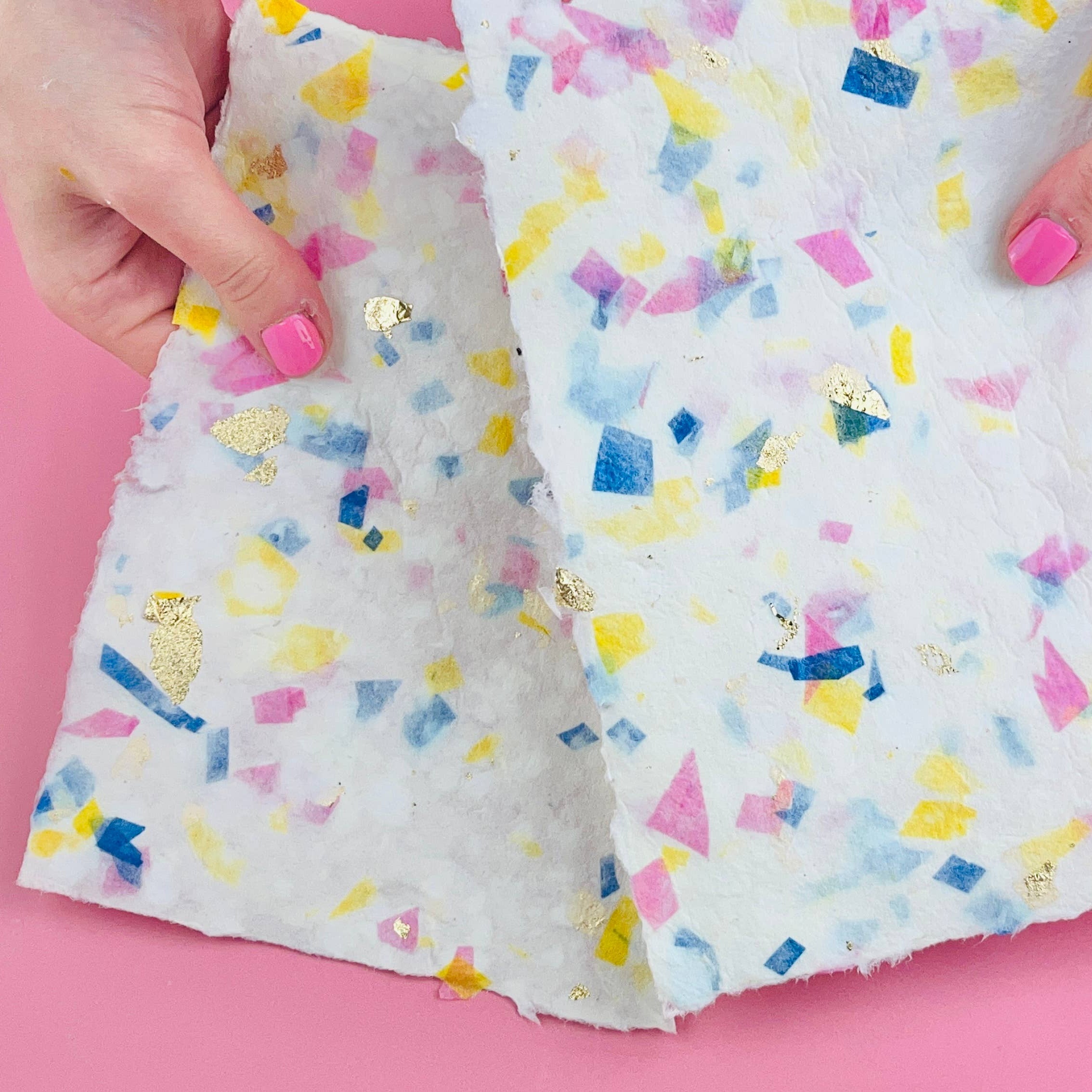 A person's hands with pink nail polish hold up a piece of white craft paper or fabric decorated with colorful confetti pieces in blue, yellow, and pink, set against a solid pink background.