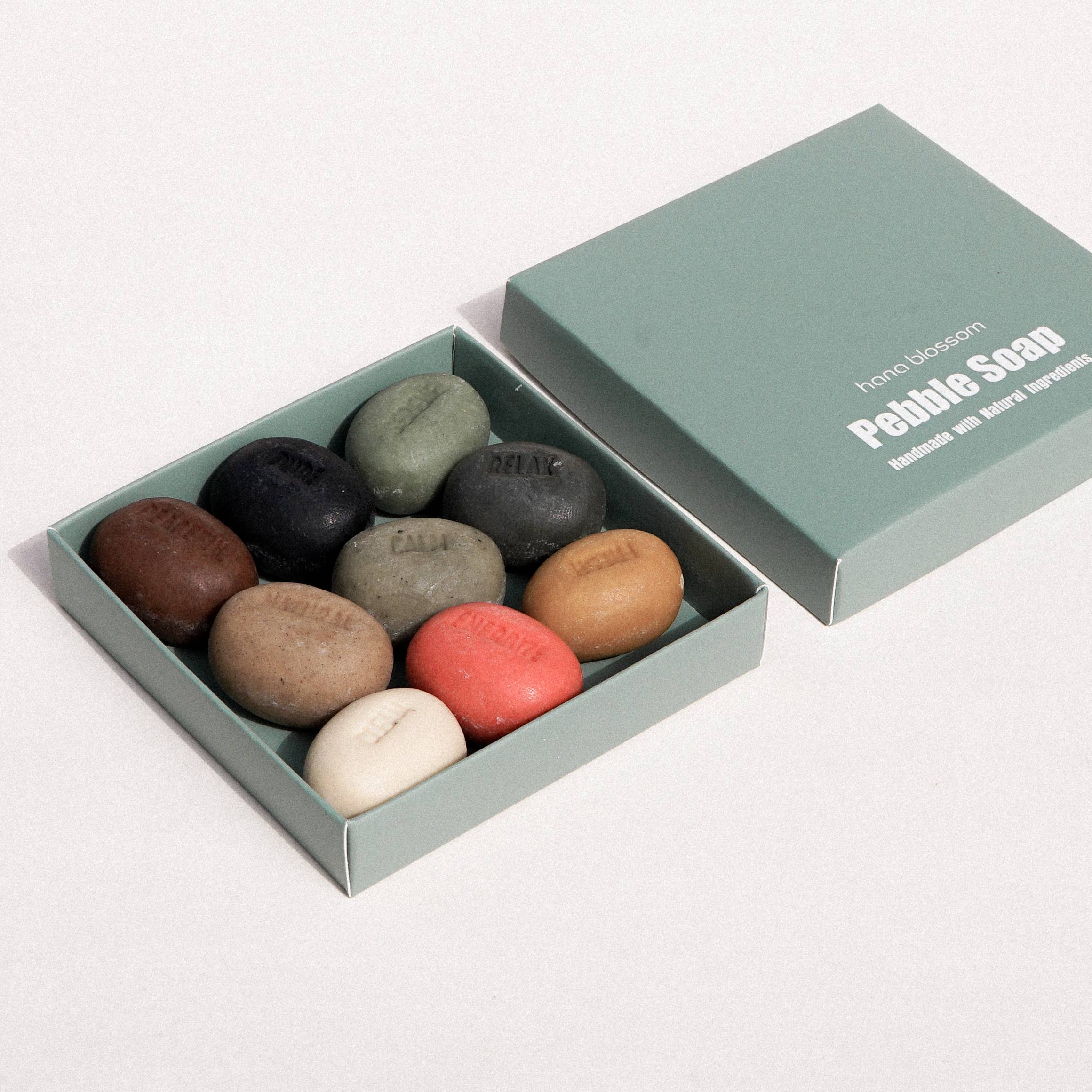 A set of ten smooth, polished pebble-shaped bath stones in various earthy colors, presented in a light sage green square box next to its lid on a clean white background. The lid features the brand name 'Pebble Soon' printed in white.