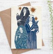 A square art print card featuring a blue and gold illustration of three women, styled on top of a brown kraft envelope over a white wood background with a green sprig.