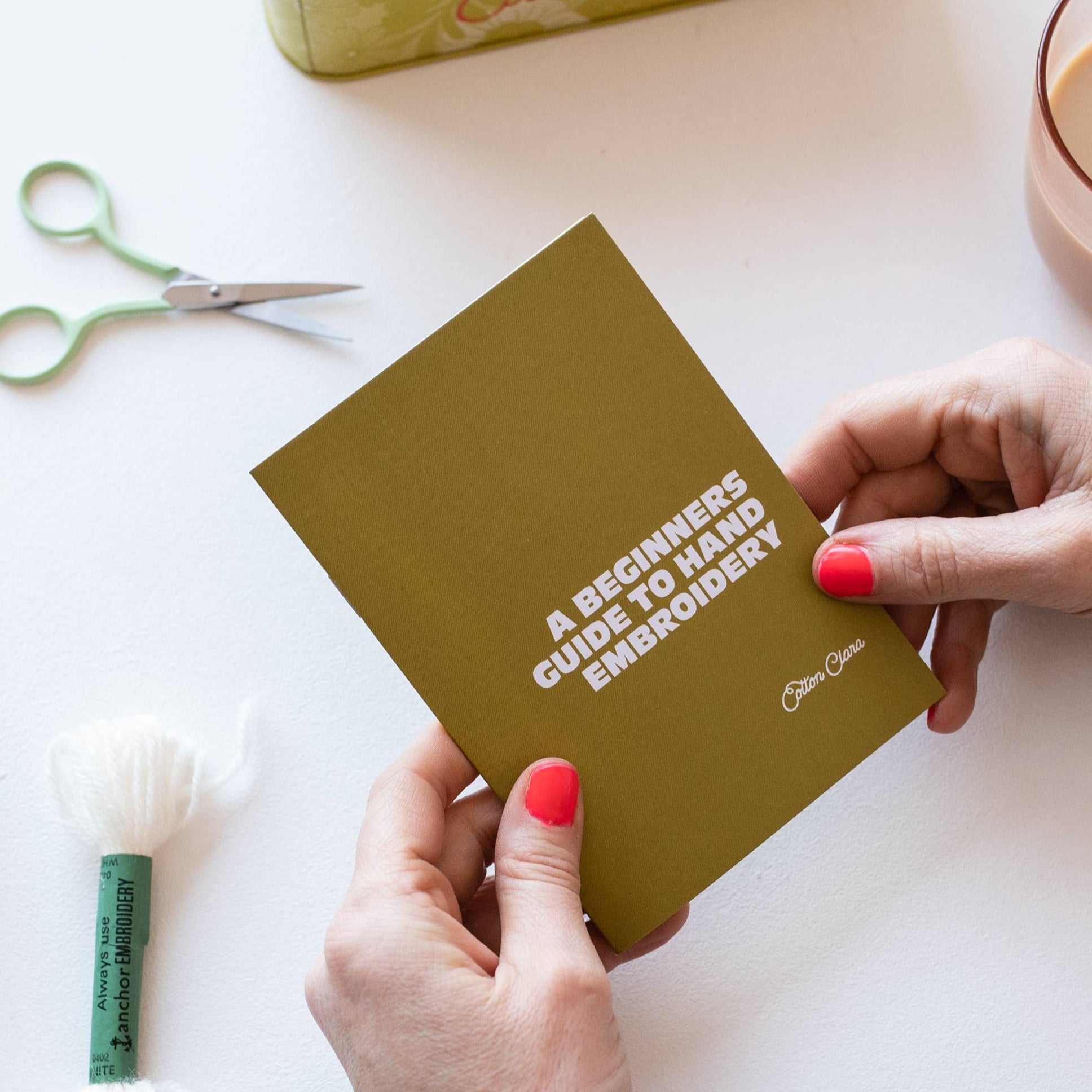 Person holding a beginners guide to hand embroidery book with craft supplies on a table.