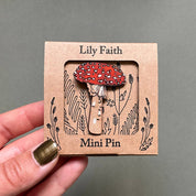 A hand with olive green nail polish holds a small square kraft paper card displaying a red and white spotted mushroom enamel pin. The card is labeled 'Lily Faith' and 'Mini Pin' with black botanical line art, set against a solid gray background.