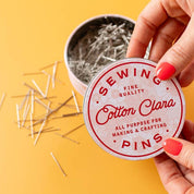 Hand holding a container of sewing pins with a yellow background