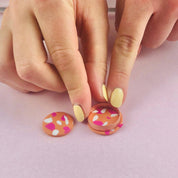 Two hands with yellow nails are placing two small round cabochons decorated with pink and white polka dots on a light lavender surface.