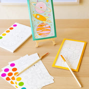 A children's painting kit displayed on a light wood table, featuring a colorful painted sample card on a small easel, white coloring pages with line art, paint-by-numbers sheets with rainbow dots, two wooden paintbrushes, and two small paint palettes.