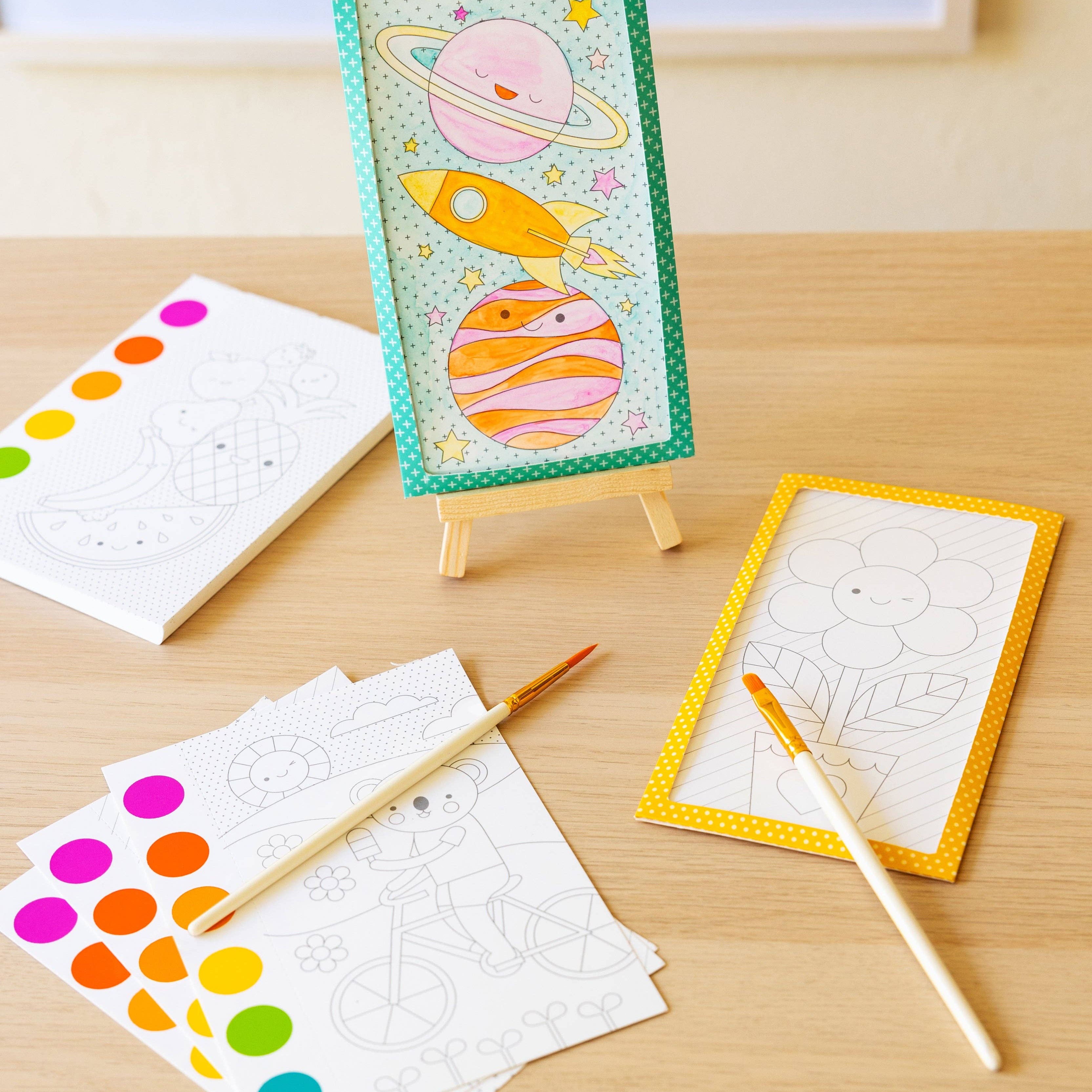 A children's painting kit displayed on a light wood table, featuring a colorful painted sample card on a small easel, white coloring pages with line art, paint-by-numbers sheets with rainbow dots, two wooden paintbrushes, and two small paint palettes.