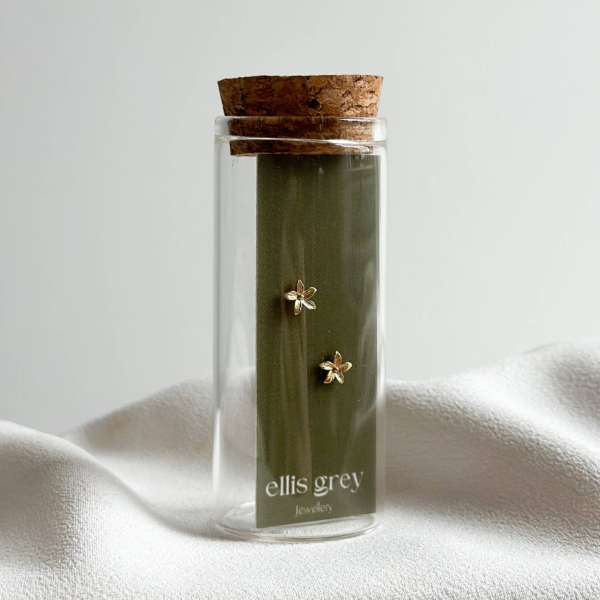 Clear glass jar with cork lid containing green tea leaves, branded 'ellis grey', on a white fabric background.