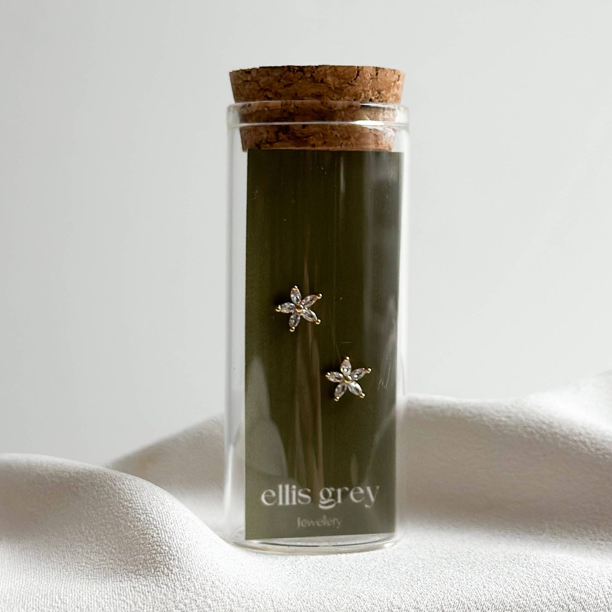 A pair of silver star-shaped stud earrings displayed inside a clear glass vial with a cork stopper, containing a dark olive green liquid. The vial is labeled 'ellis grey' and sits on draped white fabric against a light gray background.