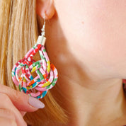A close-up lifestyle shot of a woman wearing a colorful, multi-layered knotted paper dangle earring on her ear, with her hand holding the earring to show its details against a light gray background.