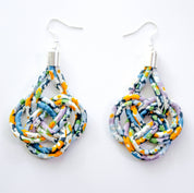 A pair of handmade teardrop-shaped dangle earrings made from knotted, braided fabric strips in a multicolor pattern of blue, orange, purple, green, and white, hanging from silver fishhook wires on a plain white background.