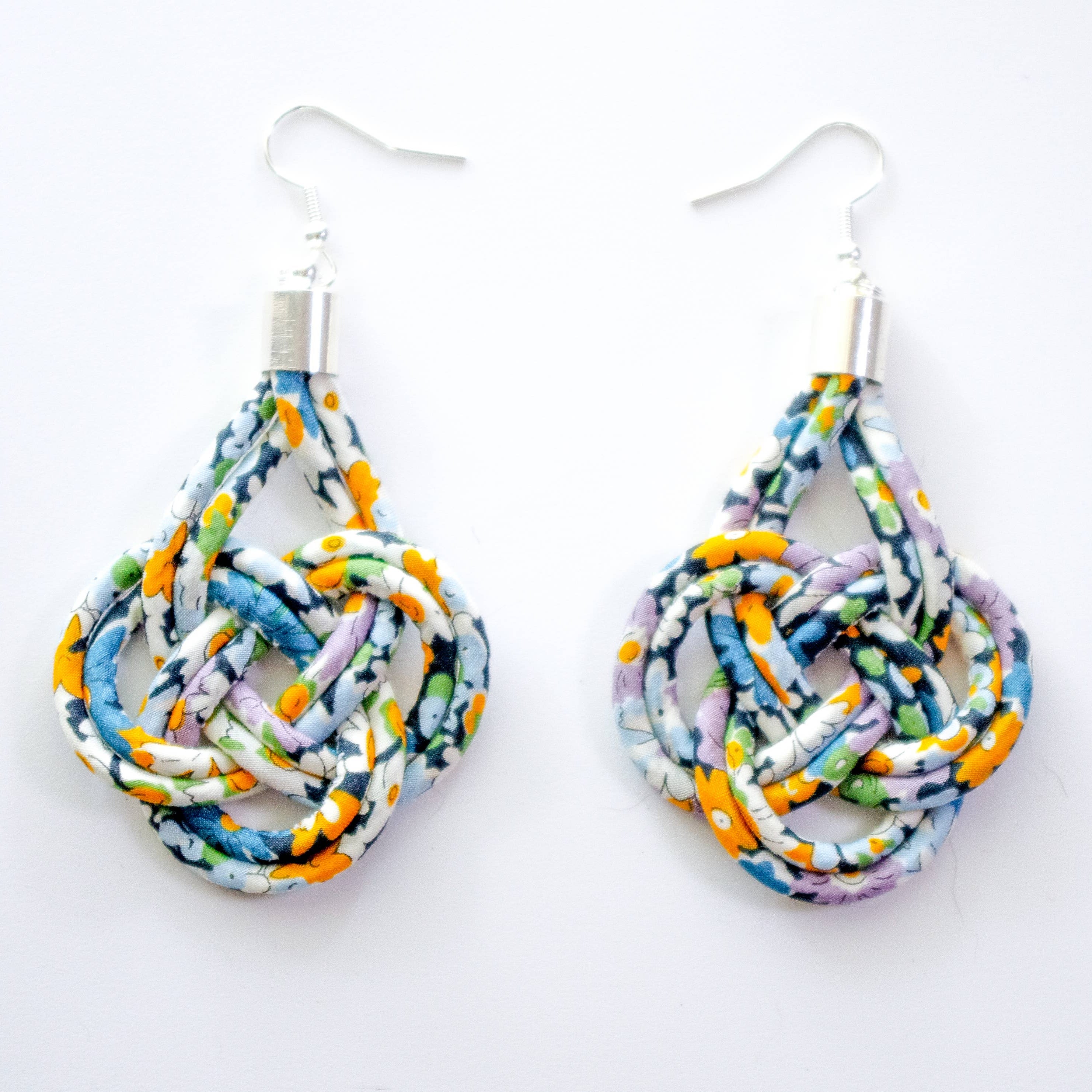 A pair of handmade teardrop-shaped dangle earrings made from knotted, braided fabric strips in a multicolor pattern of blue, orange, purple, green, and white, hanging from silver fishhook wires on a plain white background.