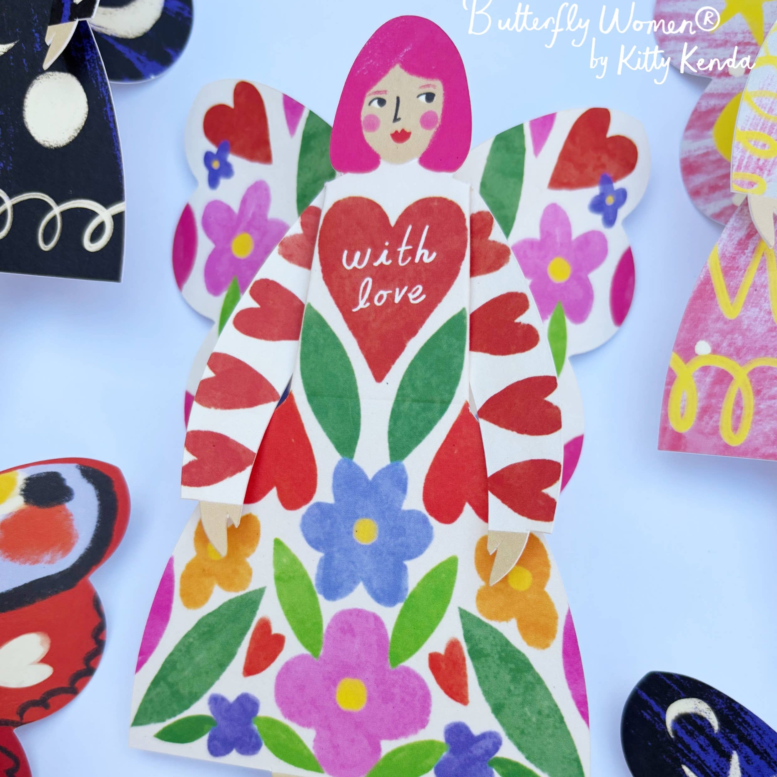 Colorful paper doll with floral design and 'with love' text, surrounded by other paper dolls on a light blue background.