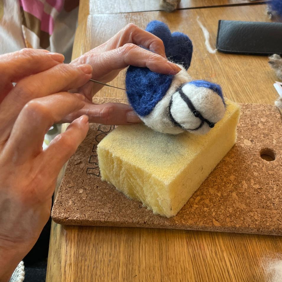Person holding a small blue and white bee toy on a wooden board with craft materials in the background.