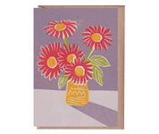 A greeting card with a linocut print of three red flowers in a yellow vase, set against a purple background, with a beige kraft envelope.