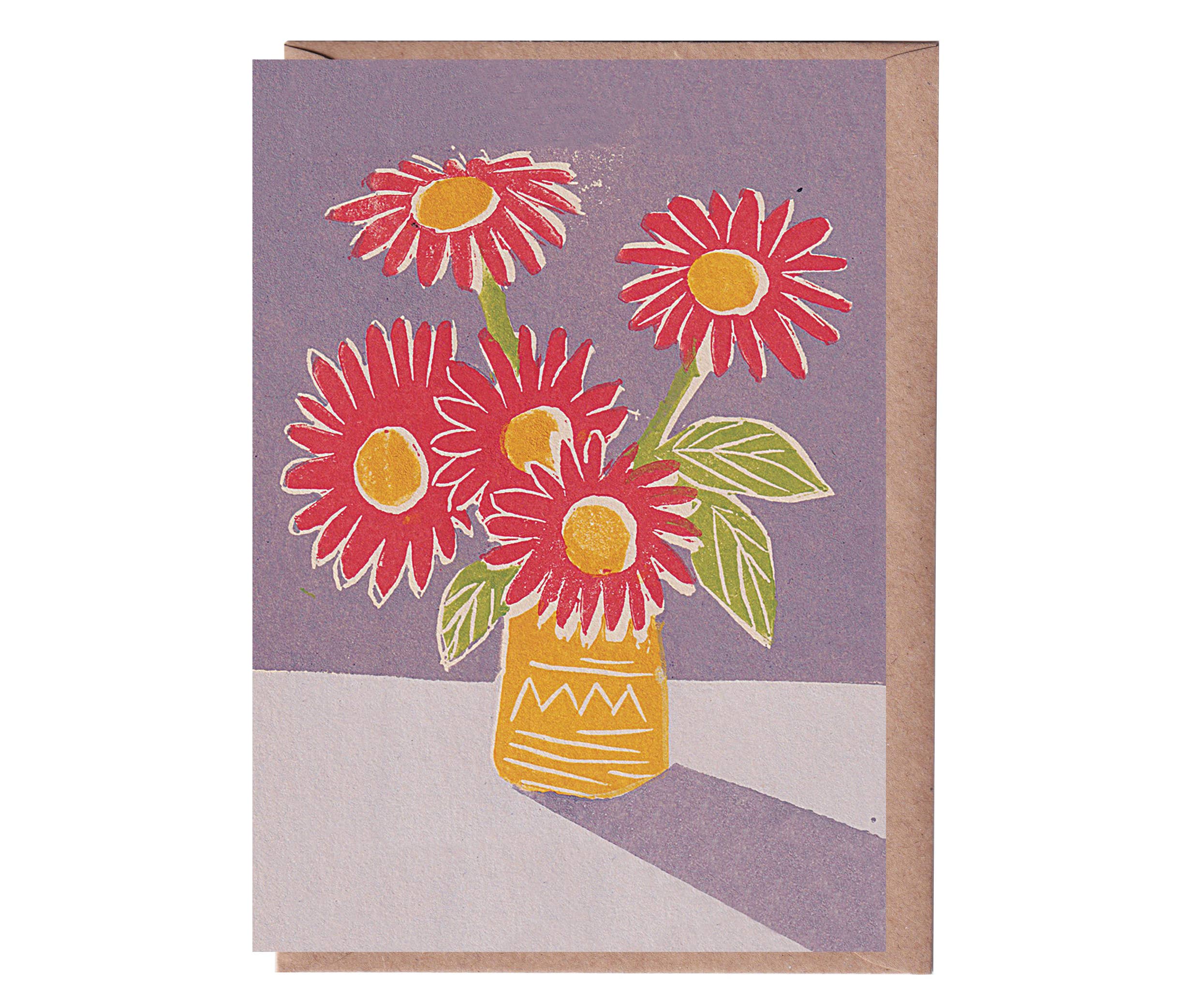 A greeting card with a linocut print of three red flowers in a yellow vase, set against a purple background, with a beige kraft envelope.