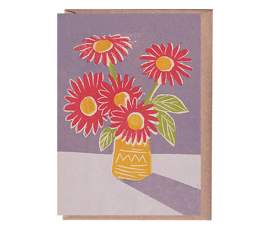 A greeting card with a linocut print of three red flowers in a yellow vase, set against a purple background, with a beige kraft envelope.