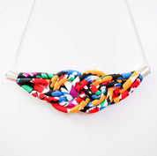 A colorful woven fabric necklace with a wide, curved band made of tightly woven multicolor threads, suspended from a white chain with silver-toned metal connectors, shown against a plain white background.