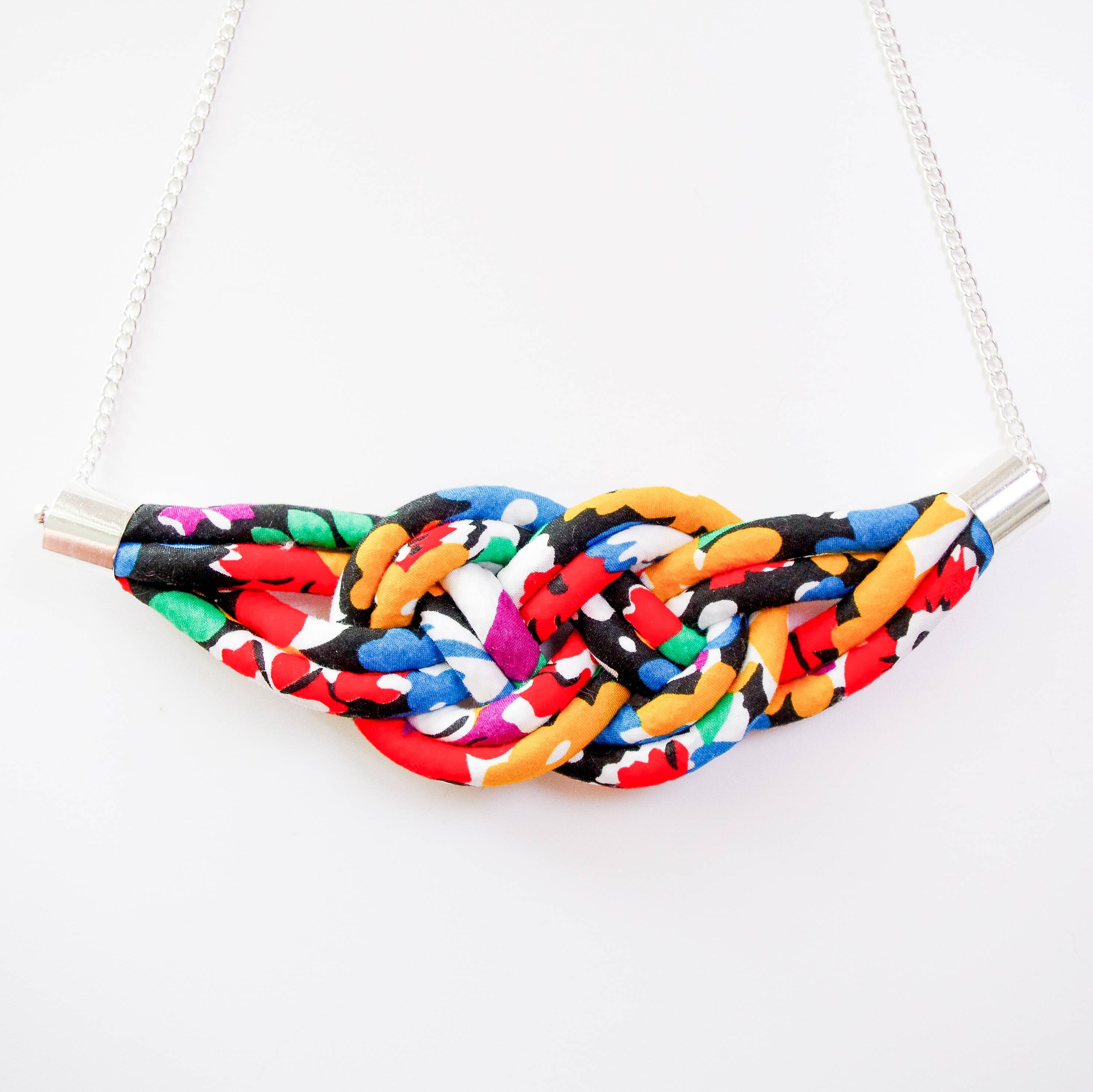 A colorful woven fabric necklace with a wide, curved band made of tightly woven multicolor threads, suspended from a white chain with silver-toned metal connectors, shown against a plain white background.