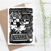Greeting card with black and white illustration of a couple kissing, envelope, and plants on a light wooden surface.