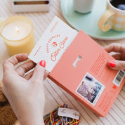 Person opening a pink box with a card on a table with a candle, cookies, and a cup of coffee.