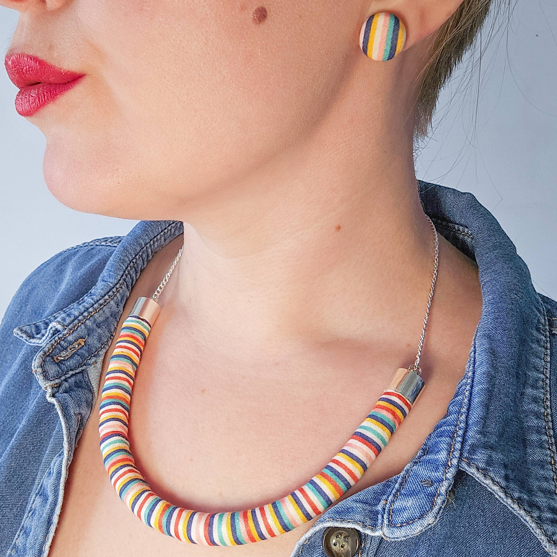 A close-up lifestyle shot of a woman wearing a chunky, multi-colored striped tube necklace and matching striped stud earrings, styled with a denim jacket.