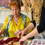 Two women working with yarn on a table, surrounded by colorful yarn balls and a pattern book.