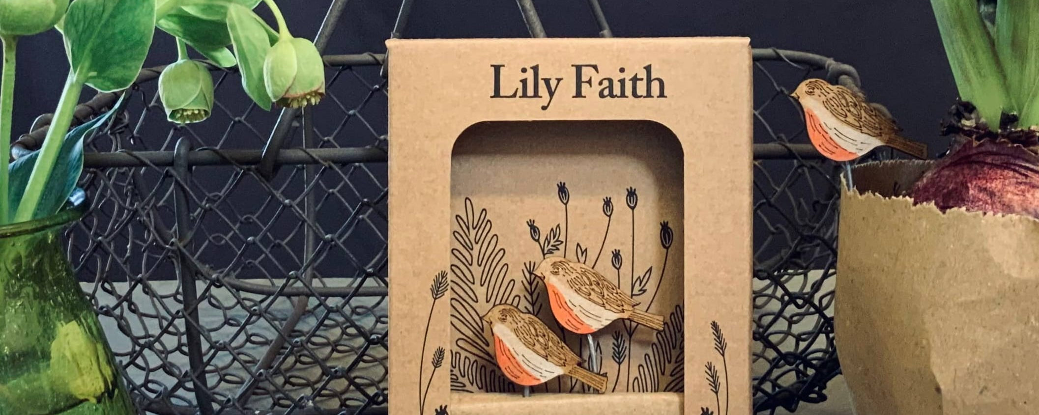A kraft paper plant pot companion box labeled 'Lily Faith' stands centered on a tabletop, surrounded by decorative plants and a green glass vase against a dark fabric backdrop.