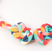 A close-up of a braided fabric strap or belt with a knot, featuring a multicolor pattern of red, orange, yellow, teal, and white on a plain white background.