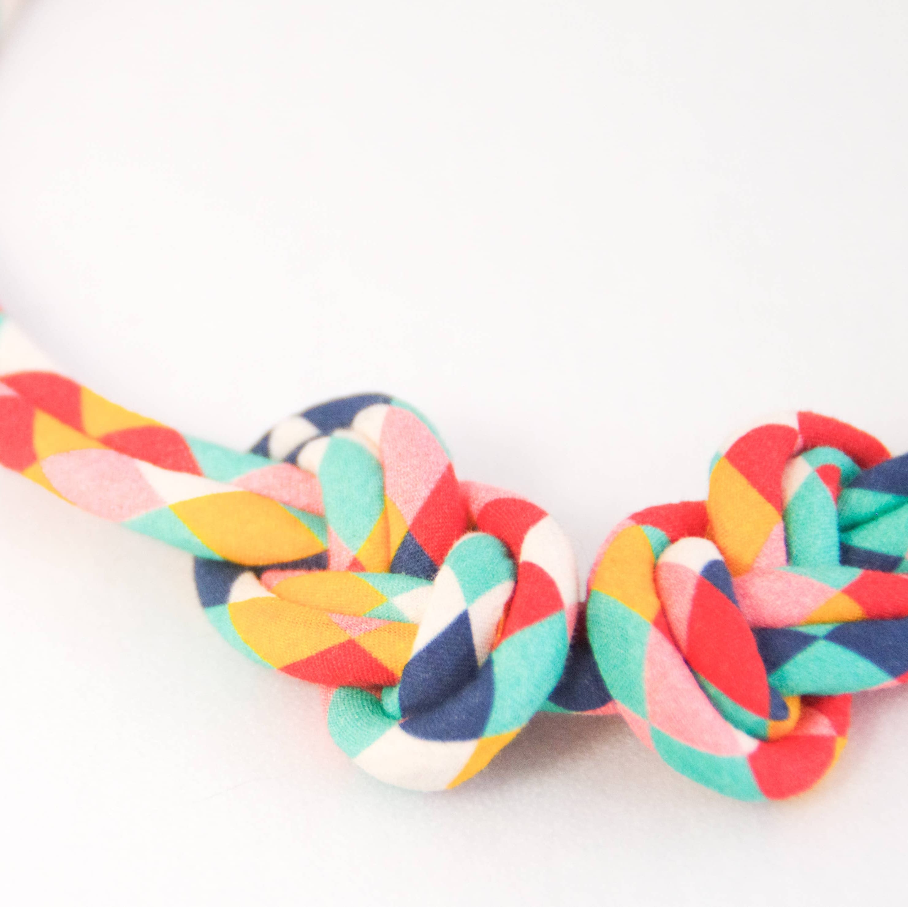A close-up of a braided fabric strap or belt with a knot, featuring a multicolor pattern of red, orange, yellow, teal, and white on a plain white background.