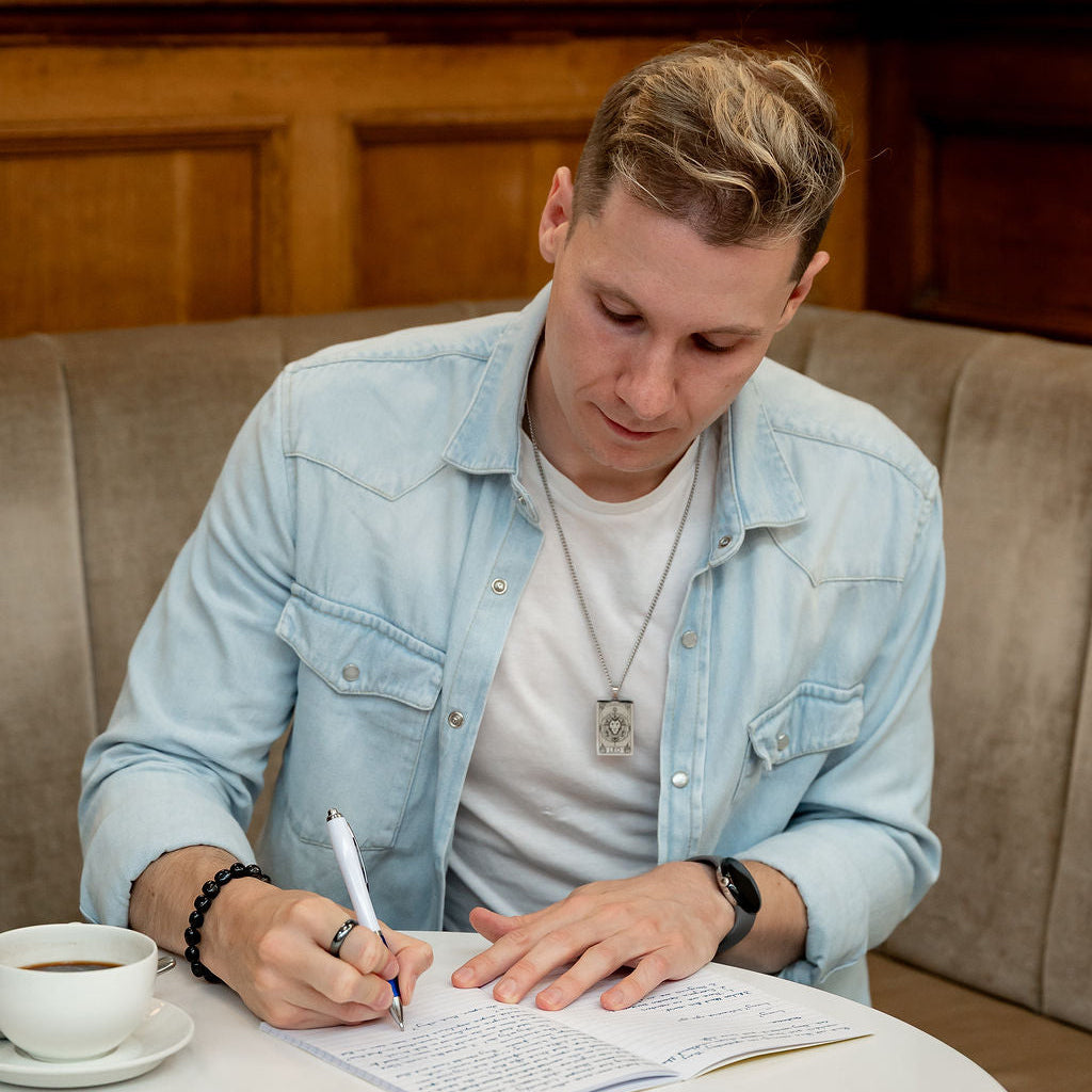 A man wearing a light blue denim jacket over a white t-shirt sits at a white table in a cafe, writing in a notebook. A cup of coffee is on the table next to him.
