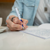 A close-up of a person's hand writing in a lined notebook with a white and blue pen, wearing a light blue denim jacket over a white shirt. A white coffee cup is visible on the left, and the background is softly blurred.