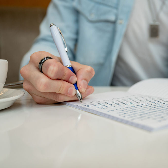 A close-up of a person's hand writing in a lined notebook with a white and blue pen, wearing a light blue denim jacket over a white shirt. A white coffee cup is visible on the left, and the background is softly blurred.