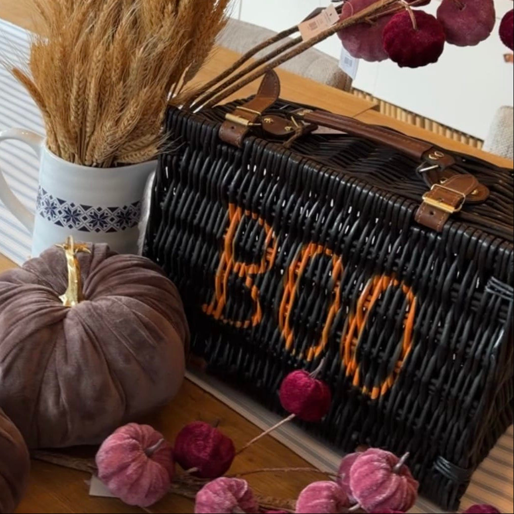A black wicker basket with gold-tone handles and the word 'BOO' in orange lettering, styled on a table with velvet pumpkins, dried wheat in a white mug, and burgundy floral stems in a shop interior.