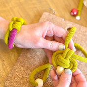 Hands tying a yellow knot with beads on a cork board, surrounded by colorful bracelets.