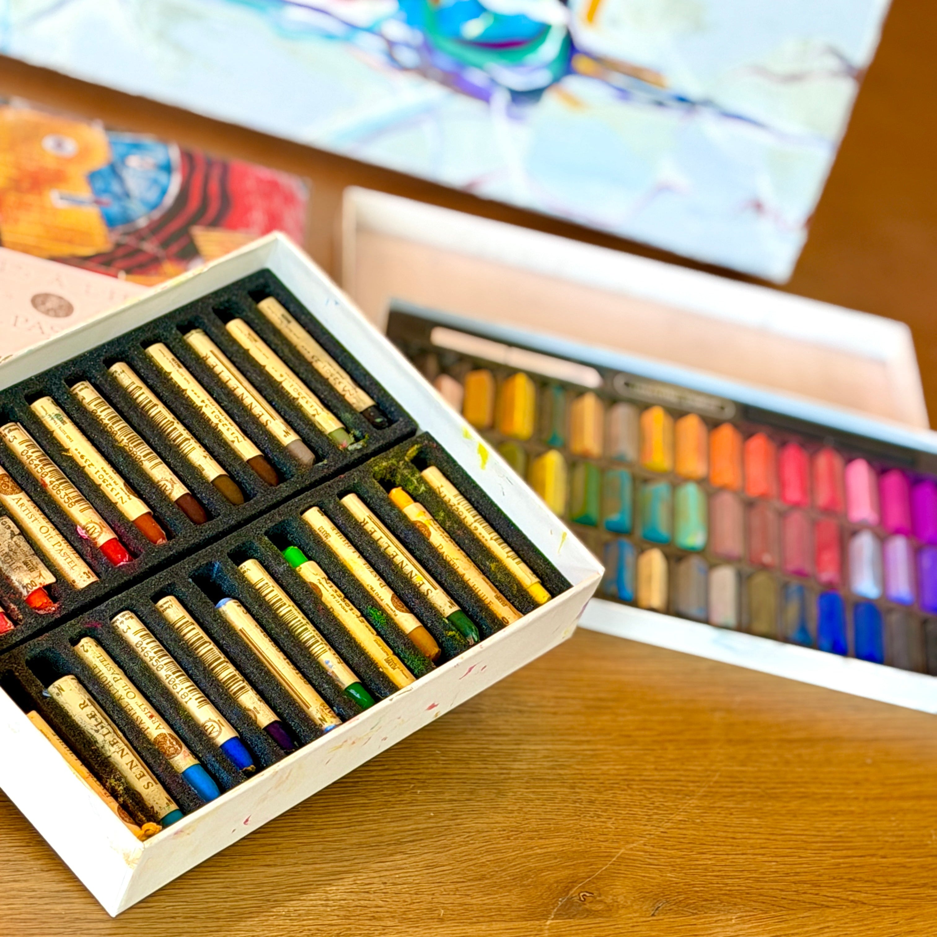 Set of pastel crayons in a wooden box with a colorful painting in the background.