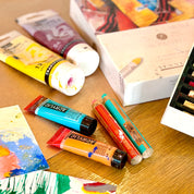 Art supplies including paint tubes and a box on a wooden surface with a painting in the background.