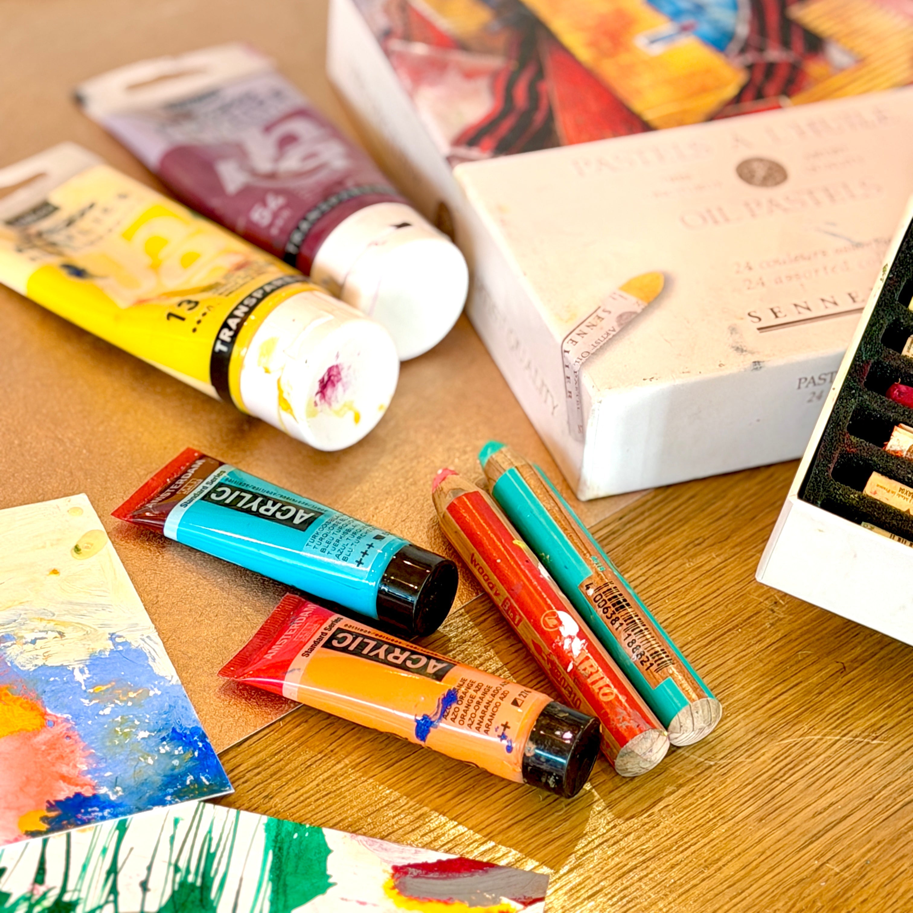 Art supplies including paint tubes and a box on a wooden surface with a painting in the background.