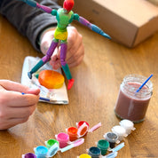 Colorful toy figure being painted with various paint colors on a wooden table.