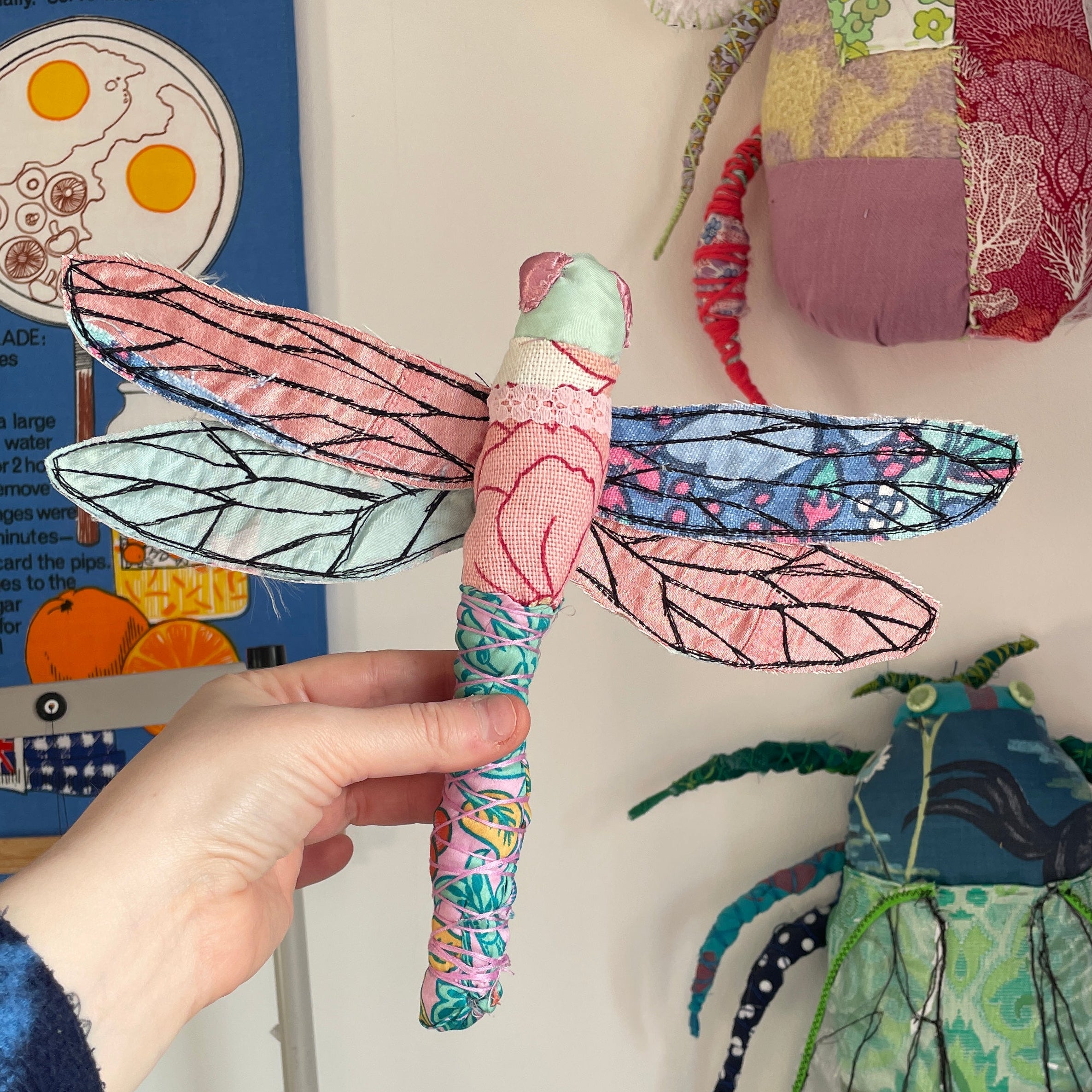 Hand holding a colorful paper dragonfly craft with fabric wings on a white surface.