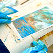 Person wearing blue gloves working on a piece of art with brushes and paint